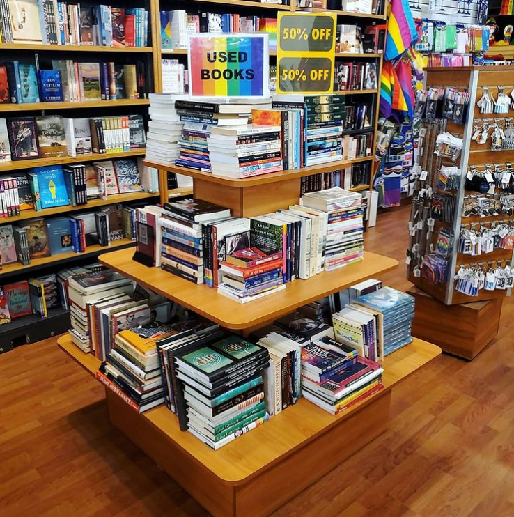 A photo of the interor of the Little Sister's store. A display of used books on a tired table with signs denoting 50% off. In the background there are more books and bookshelves along with rainbow flags.