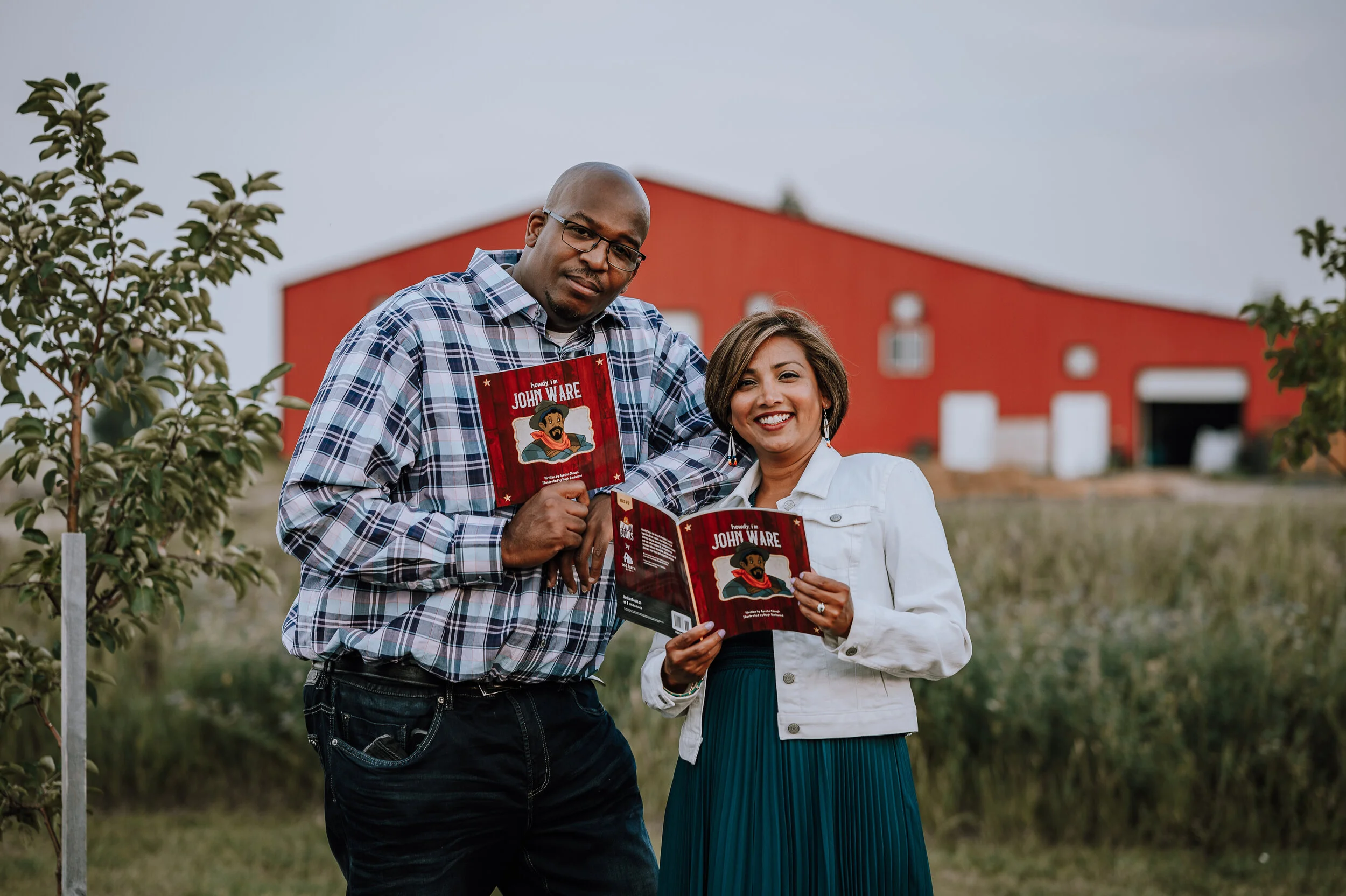 Photo of Hugh Rookwood and Ayesha Clough each holding a copy of Howdy I'm John Ware, with a red barn in the background.