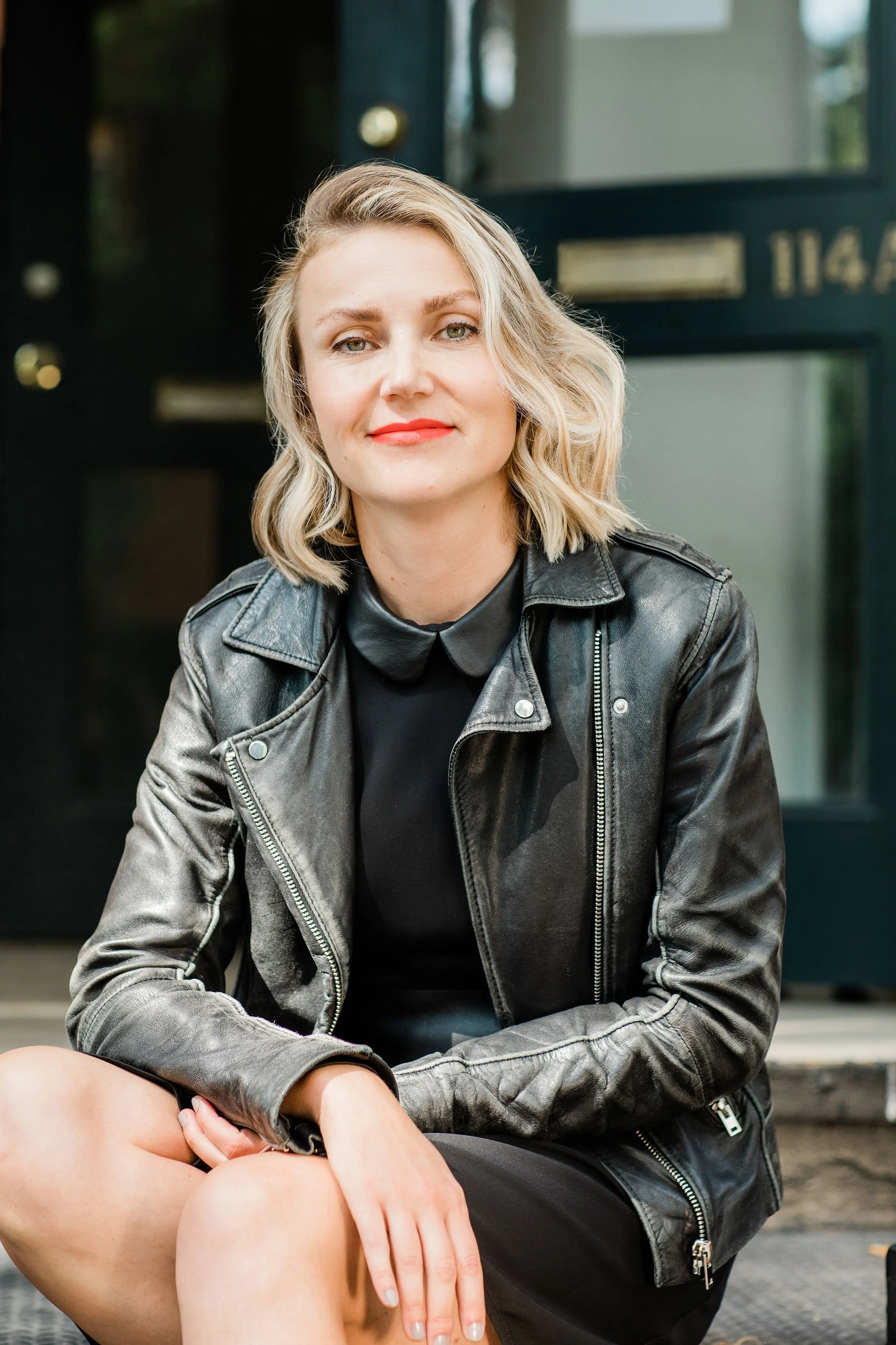 Headshot of Eva Jurczyk, Eva has blonde hair, she's wearing a black dress and a black leather jacket, She's looking at the camera and smiling.